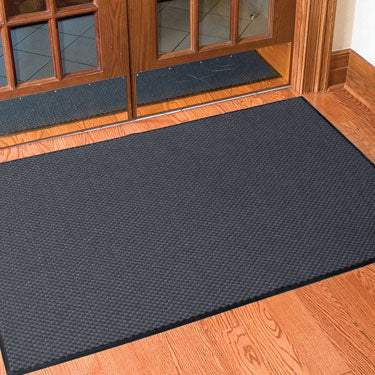 Entrance mat in front of double glass doors on wood flooring, black ribbed mat capturing dirt and moisture.