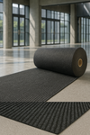 Rolled heavy-duty anthracite entrance mat unrolled on polished lobby floor with glass walls; ribbed surface channels dirt and moisture and protects flooring in busy commercial buildings