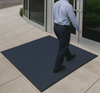 Blue heavy-duty entrance mat installed outside glass doors with person walking across; textured surface absorbs dirt and moisture and provides non-slip traction; protects entrance flooring in busy commercial spaces.