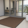 Brown moisture-trapping entrance mat inside modern office entryway with glass doors and plant; durable olefin pile absorbs dirt and moisture; ribbed vinyl backing keeps mat in place, reduces slips and protects floors.