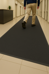 Heavy-duty non-slip rubber runner in office corridor with person walking away; long ribbed mat channels water, reduces slips, and protects polished tile floor.