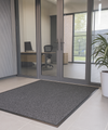 Grey moisture-trapping entrance mat with mottled design in modern office entryway with glass doors, plant and chair; durable olefin pile absorbs dirt and moisture; low-profile ribbed vinyl backing keeps mat in place, reduces slips and protects floors.
