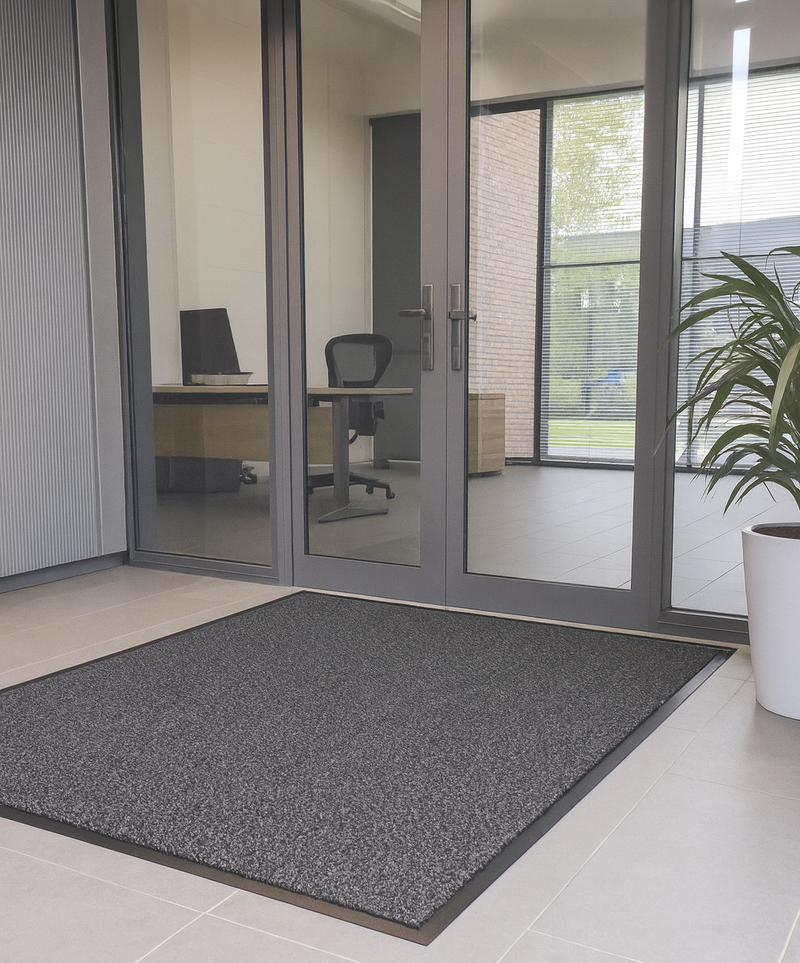 Grey moisture-trapping entrance mat with mottled design in modern office entryway with glass doors, plant and chair; durable olefin pile absorbs dirt and moisture; low-profile ribbed vinyl backing keeps mat in place, reduces slips and protects floors.