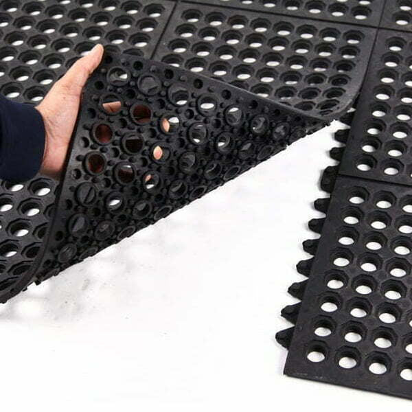 Hands lifting corner of Slip-Not hollow ring interlocking drainage rubber mat tile; close-up of open ring matting showing ring holes and interlocking edges, heavy-duty natural rubber tile measuring 0.91 m x 0.91 m and 15 mm thick for drainage, anti-slip and anti-fatigue support