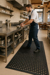 Worker moving pot on Slip-Not workstation rubber matting in a commercial kitchen; black anti-fatigue rubber mat with open-circle holes and bevelled edges provides cushioning, drainage and non-slip traction. Mat measures 1.52 m x 0.91 m and 12.5 mm thick.