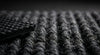Extreme close-up of anthracite commercial entrance matting pile — cut-and-loop polypropylene texture, rubber waffle backing detail, dark charcoal grey fibres