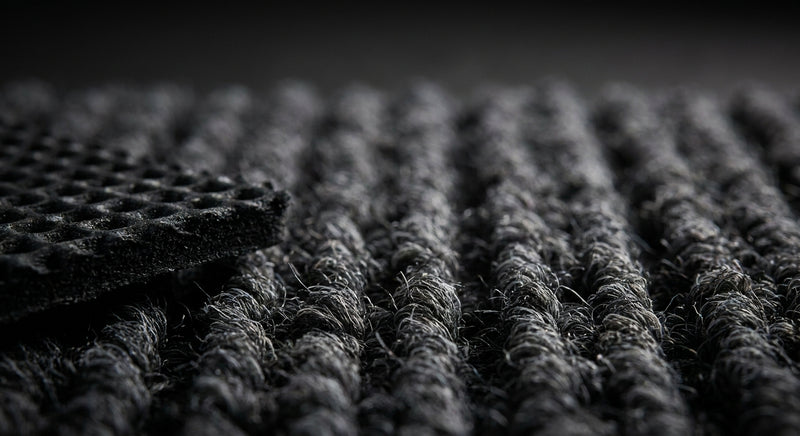 Extreme close-up of anthracite commercial entrance matting pile — cut-and-loop polypropylene texture, rubber waffle backing detail, dark charcoal grey fibres
