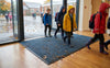 Blue waffle entrance mat at UK school entrance — autumn rainy day, children stepping in, blue mat absorbing moisture and trapping debris
