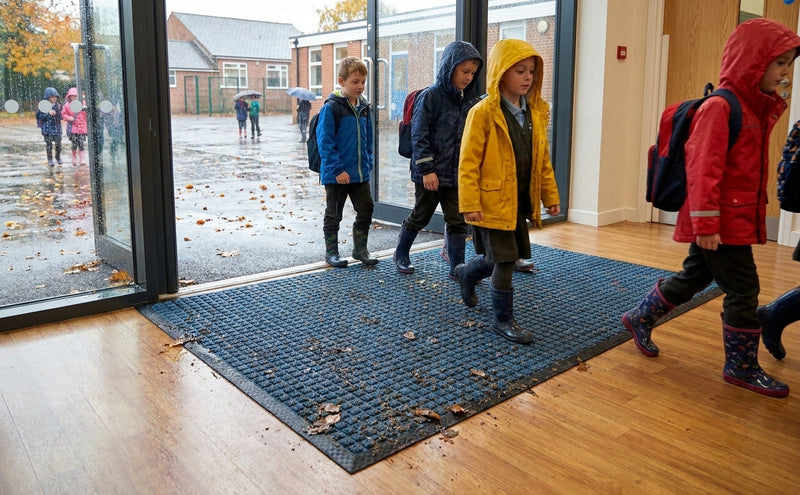 Blue waffle entrance mat at UK school entrance — autumn rainy day, children stepping in, blue mat absorbing moisture and trapping debris