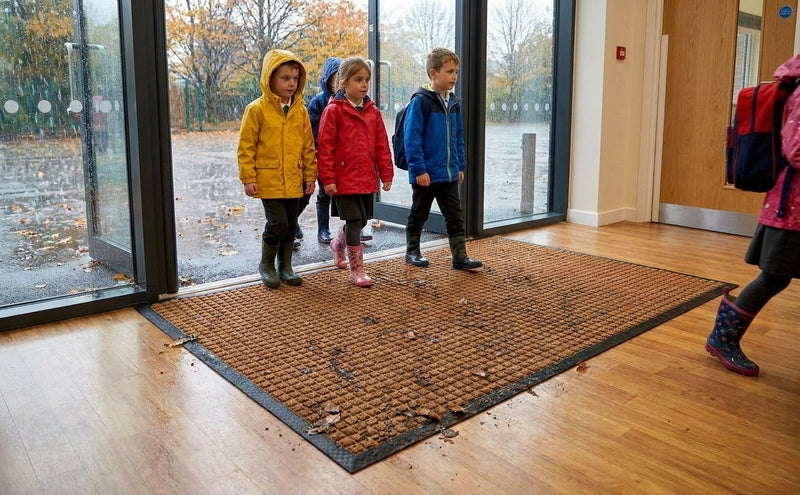Brown waffle absorbent entrance mat at UK primary school — wet rainy entrance, children with muddy boots, waffle grid surface working
