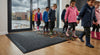 Charcoal grey waffle entrance mat at UK primary school — children in raincoats walking in from rainy day, mat trapping mud and moisture
