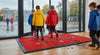 Red waffle entrance mat at UK school rainy day — bright red mat at school entrance, children in raincoats, mud and leaves visible, mat performing