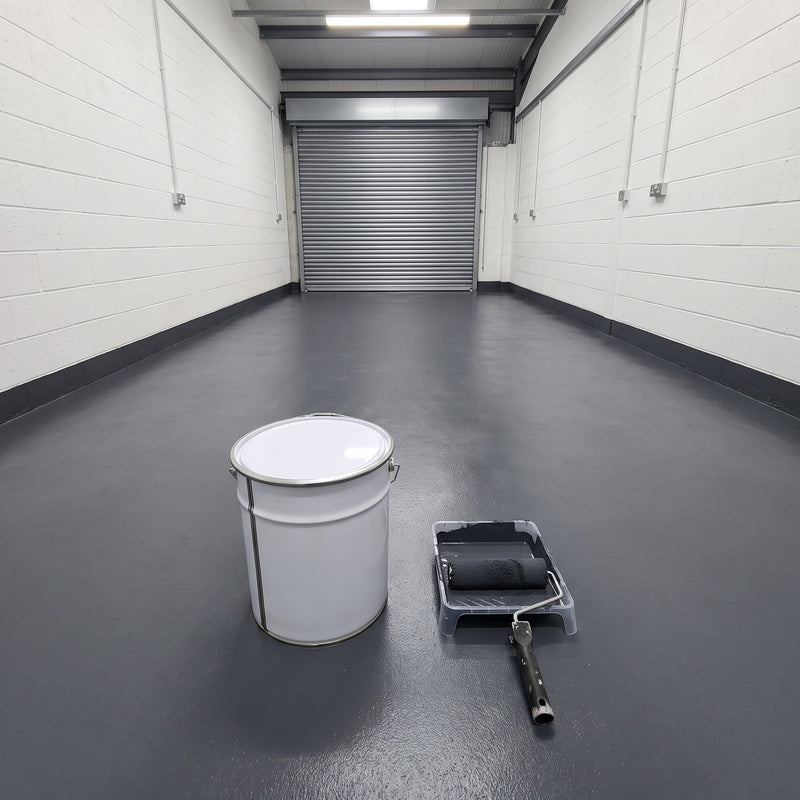 Dark grey floor paint in an empty vehicle workshop bay with a roller shutter door, 20 litre tin and roller