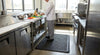 Black anti-fatigue mat at commercial kitchen workstation — 9mm foam, ribbed texture, chef standing during food preparation