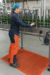 Worker standing on oil-resistant nitrile anti-fatigue rubber mat in bottling plant; orange perforated mat with drainage holes and beveled edges providing slip-resistant cushioning and comfort, measuring 1.5 m x 0.9 m and 12.5 mm thick, ideal for industrial workstations.
