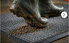 Charcoal waffle absorbent mat — muddy Wellington boots stepping onto mat, waffle grid scraping mud and trapping moisture from boot soles, black rubber border