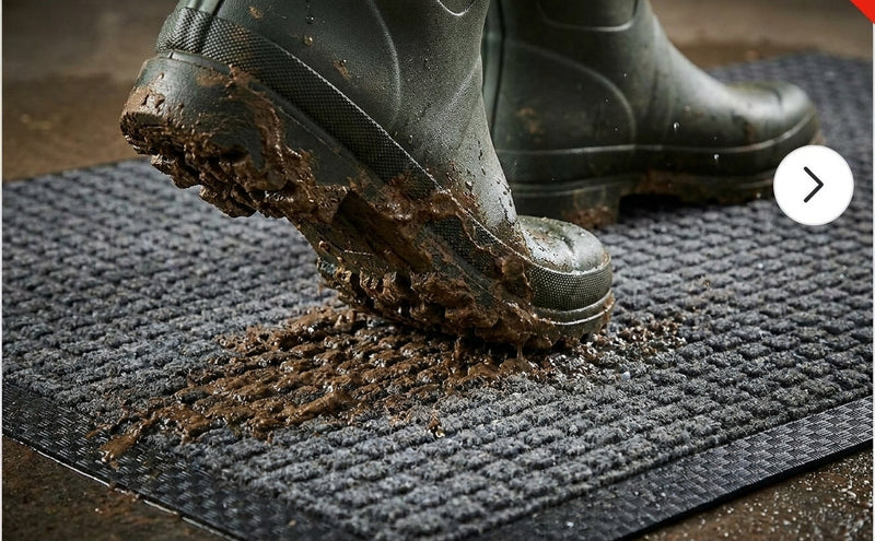 Charcoal waffle absorbent mat — muddy Wellington boots stepping onto mat, waffle grid scraping mud and trapping moisture from boot soles, black rubber border