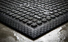 Close-up corner detail of charcoal waffle absorbent mat — raised waffle pile absorbing moisture, black criss-cross rubber border, low angle dramatic lighting