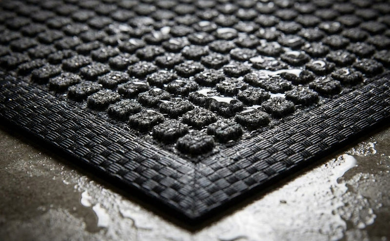 Close-up corner detail of charcoal waffle absorbent mat — raised waffle pile absorbing moisture, black criss-cross rubber border, low angle dramatic lighting