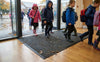 Waffle absorbent entrance mat at UK primary school rainy day — children in wet coats stepping on charcoal mat, clean dry floor visible beyond