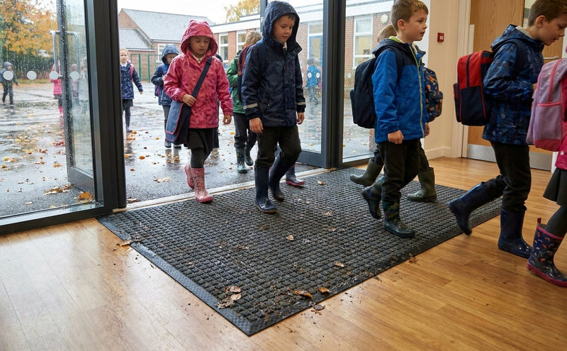 Waffle absorbent entrance mat at UK primary school rainy day — children in wet coats stepping on charcoal mat, clean dry floor visible beyond