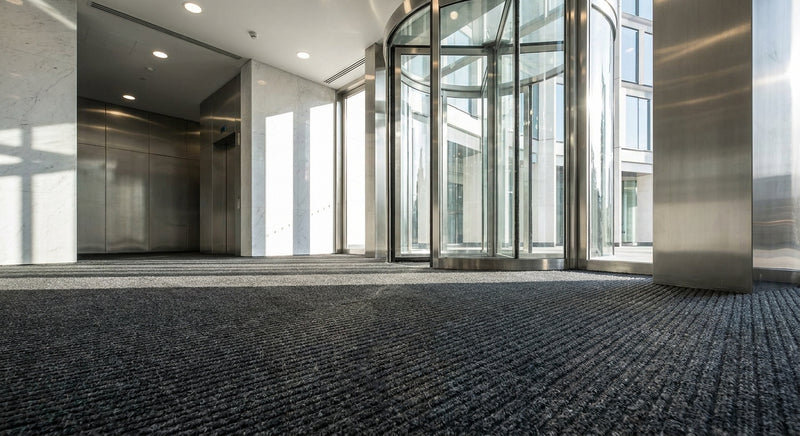 Anthracite ribbed entrance carpet matting in luxury corporate building with glass revolving door — premium dark grey polypropylene cut-and-loop pile, 12mm thick, rubber waffle backed
